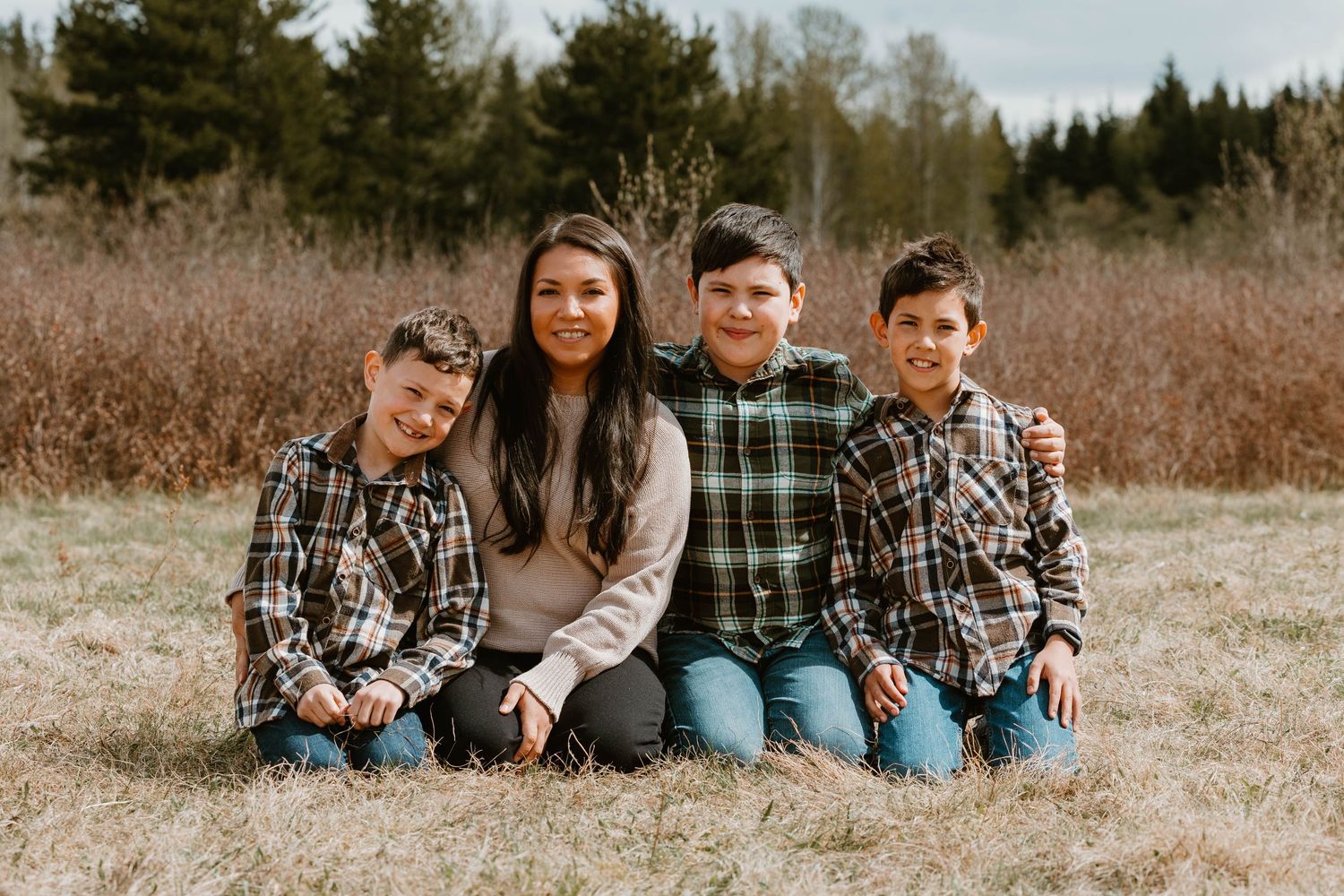 A casual outdoor family portrait taken in a field with pine trees in the background.