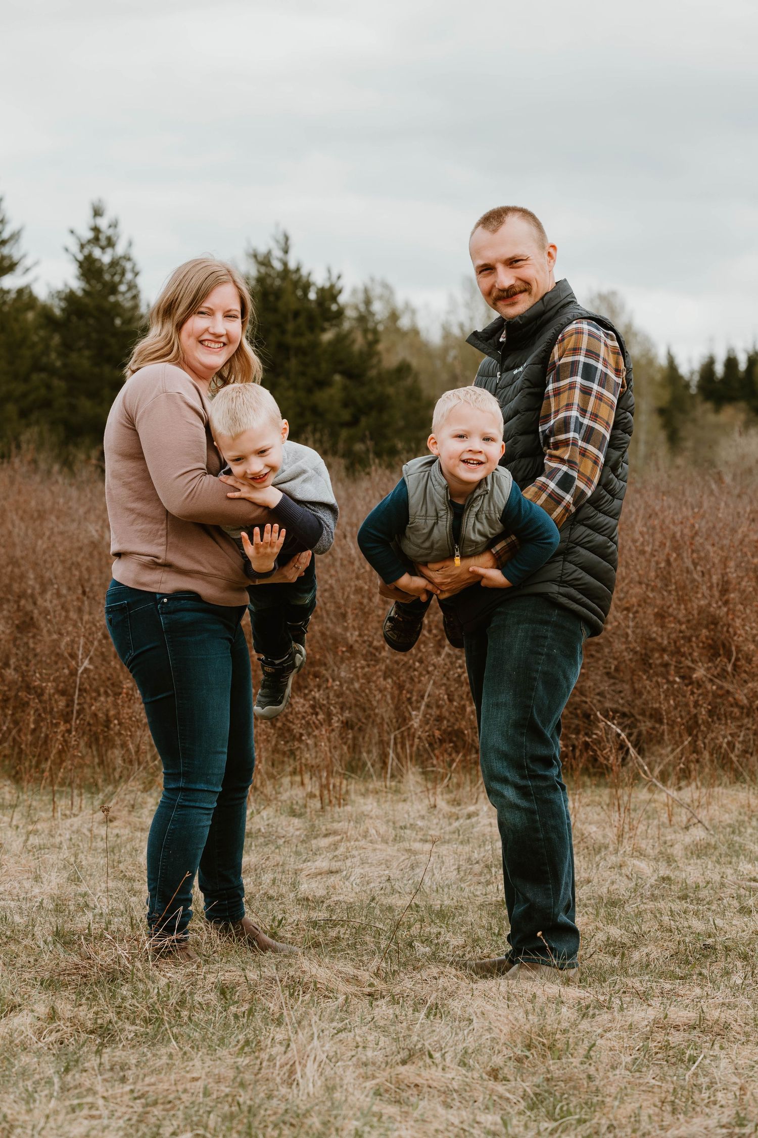 Family portrait in casual attire standing together in field with evergreen backdrop.