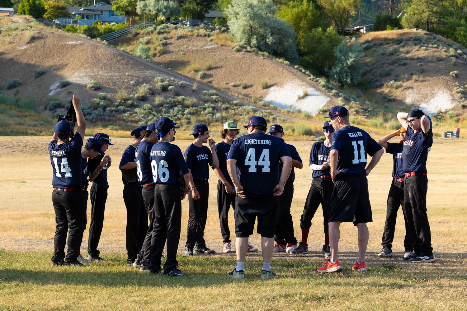 Baseball team huddles together in navy uniforms on a sunlit field for a team meeting.