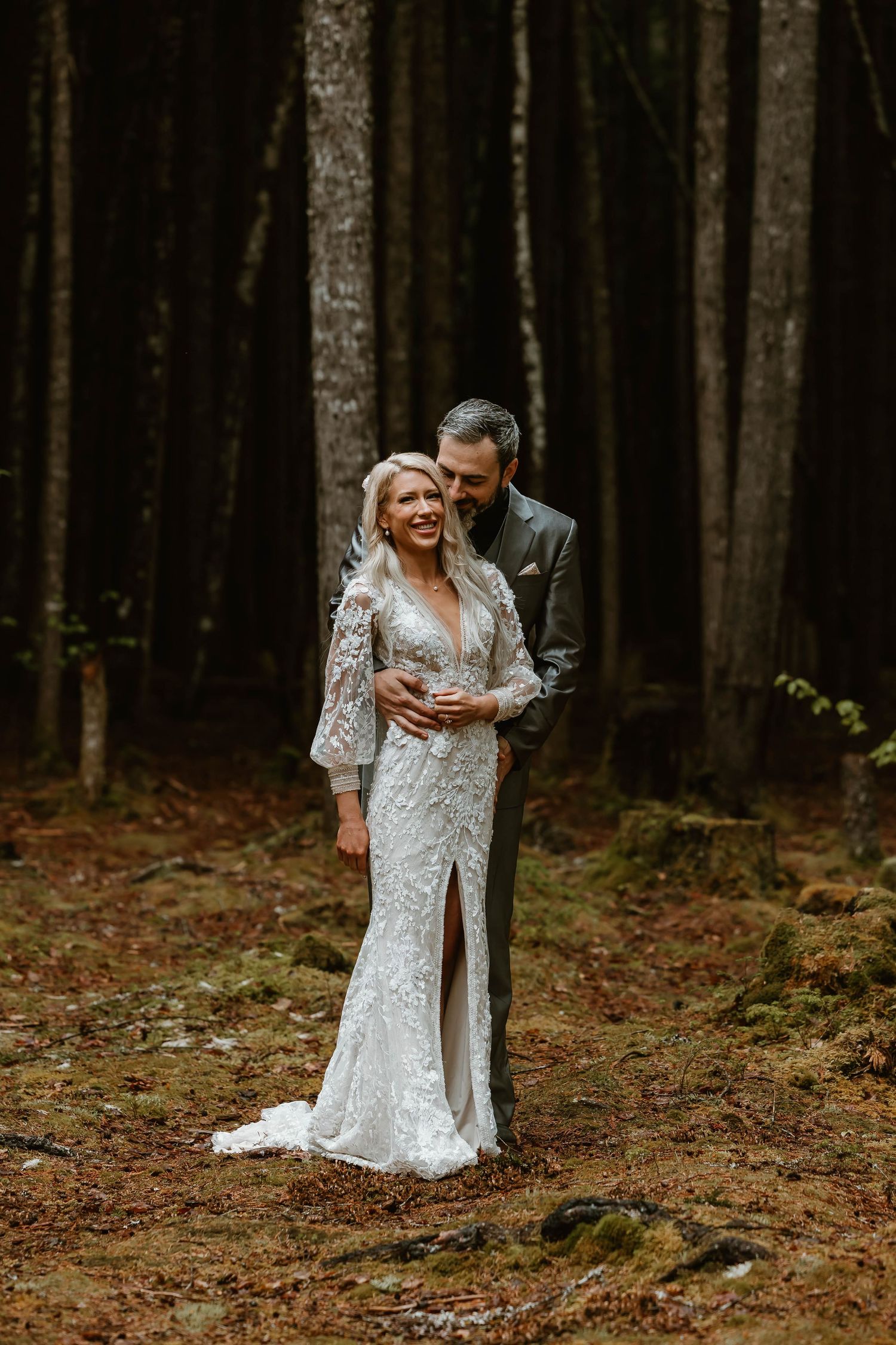 A romantic wedding portrait in a pine forest with a couple embracing, the bride in a fitted lace gown.