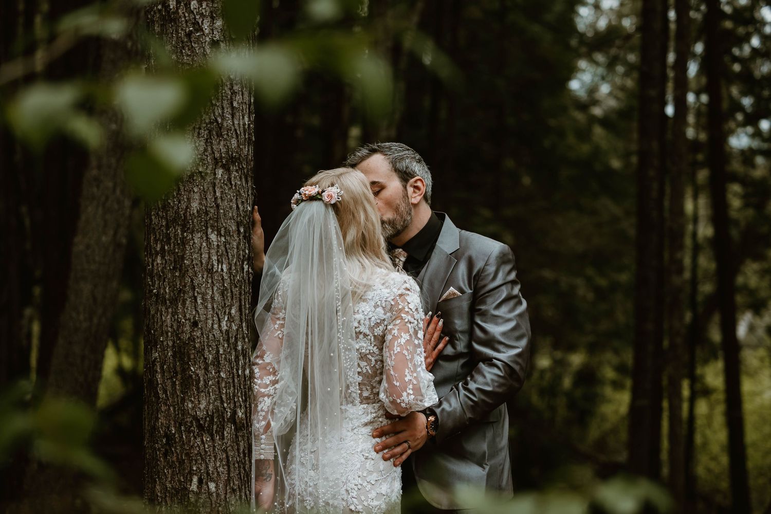 A romantic wedding couple embraces against a tree in a moody forest setting wearing elegant attire with a flowing veil.