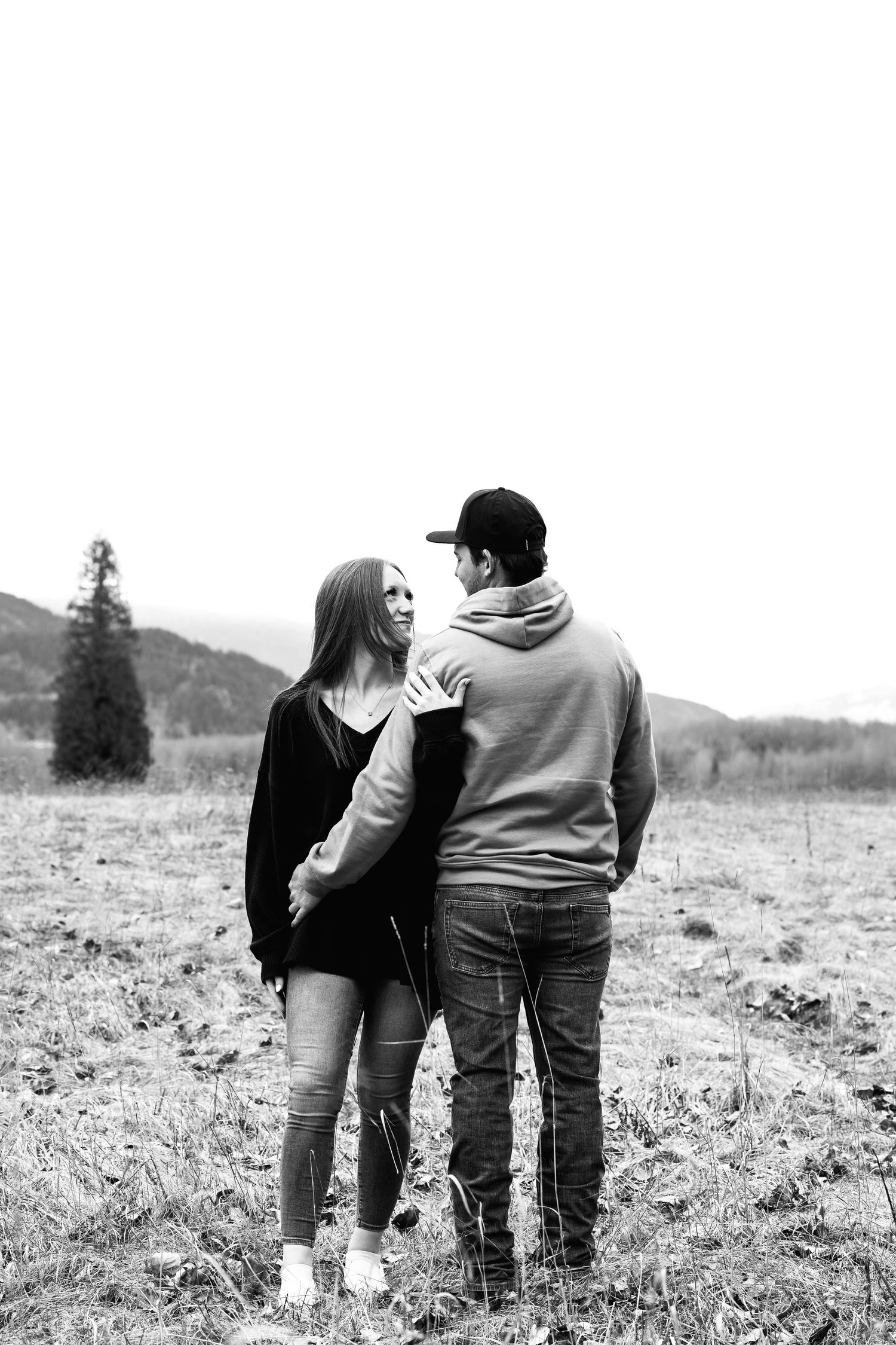 Couple shares intimate moment while standing in field with mountain backdrop in black and white photograph.