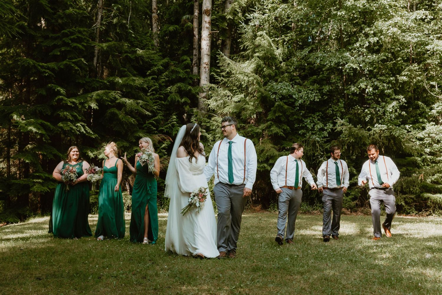 Wedding party in green dresses and suspenders poses in forest setting.