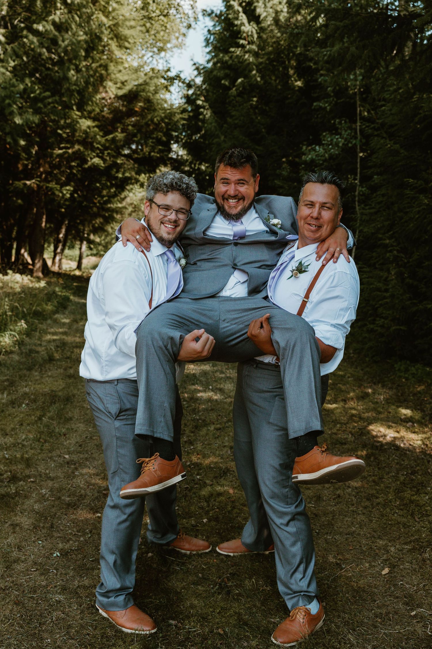 Groomsmen in suspenders playfully carrying groom in grey suit outdoors.