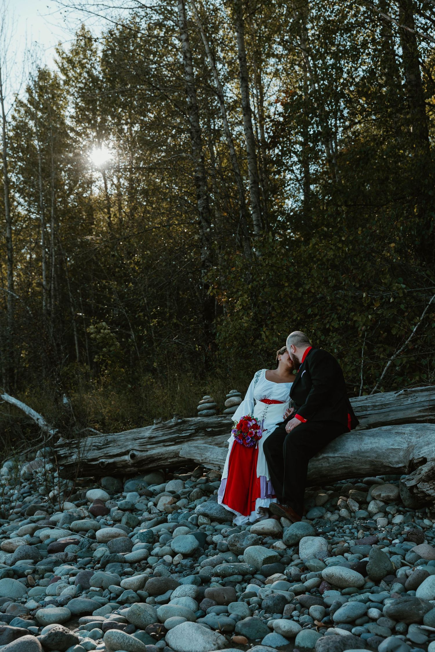 Couple sits on rocky shore against sunlit forest backdrop.