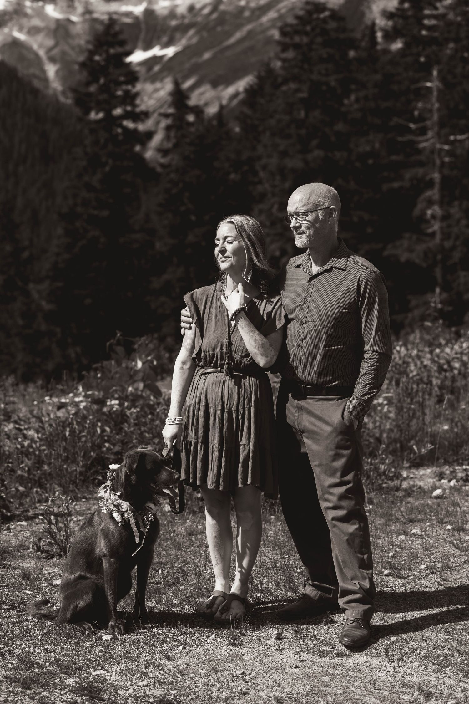 A couple and their chocolate lab dog pose for a black and white portrait in a mountainous landscape.
