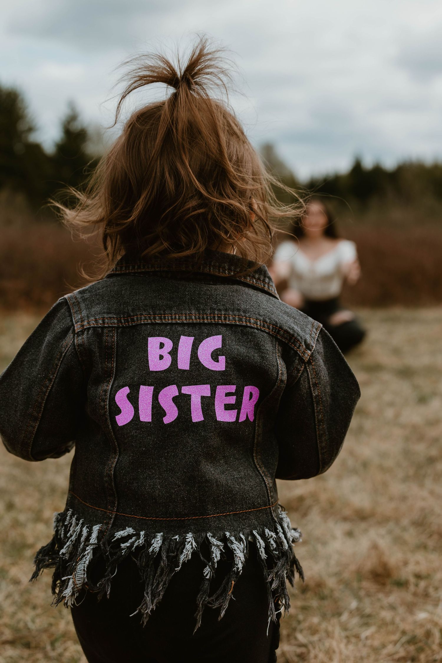 A denim jacket with purple 'Big Sister' text seen from behind in an outdoor field setting.
