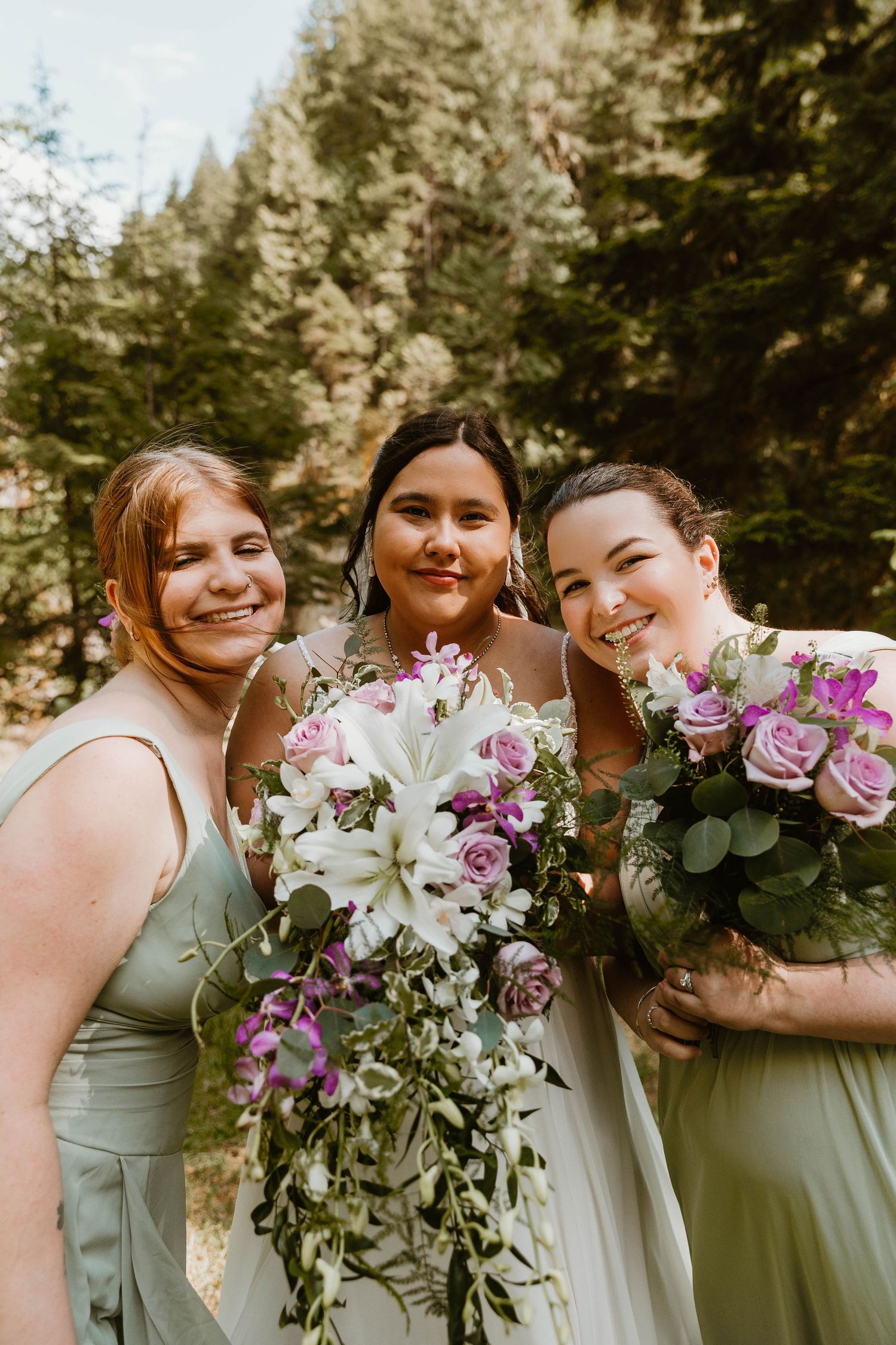Three women smile while holding floral bouquets featuring purple and white flowers.