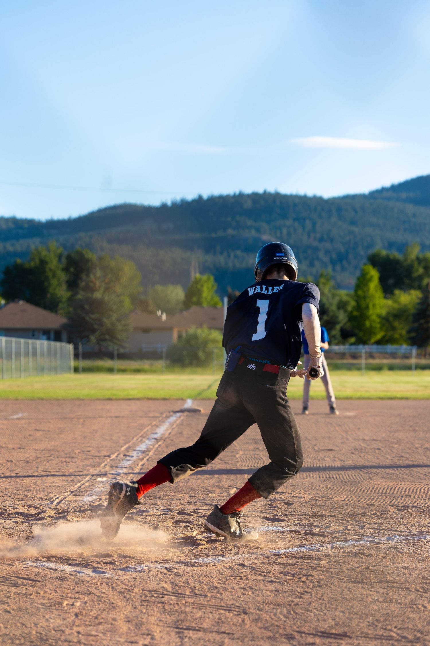 Baseball player in black uniform sliding into base creating dust cloud on dirt field with mountains in background.
