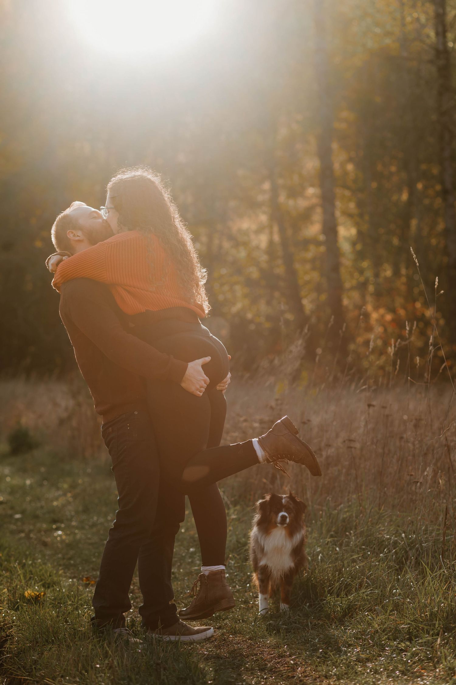 A couple embraces in golden sunset light while their Australian Shepherd dog stands nearby.