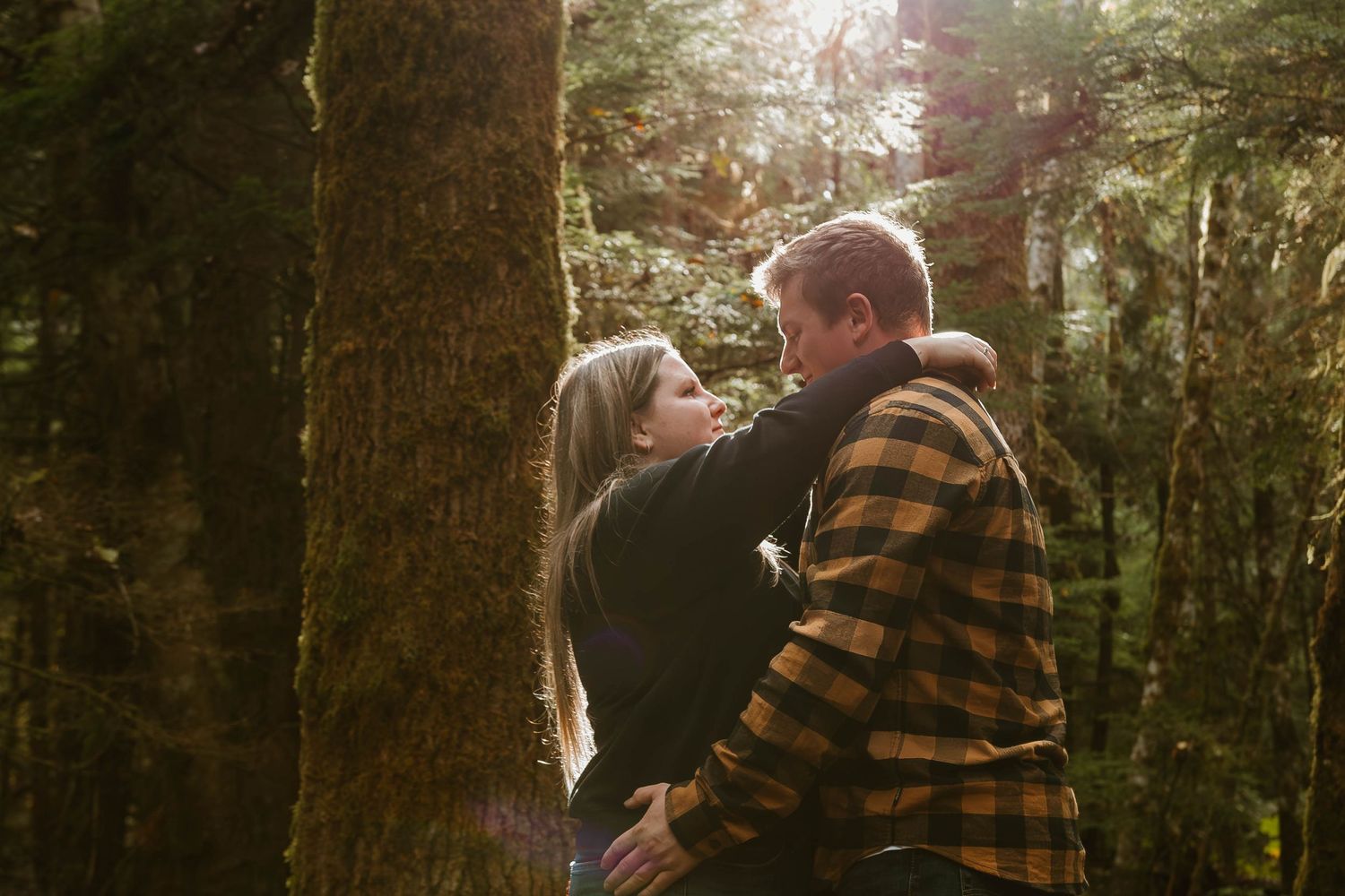 A couple in casual attire embracing near large trees in a forest setting.