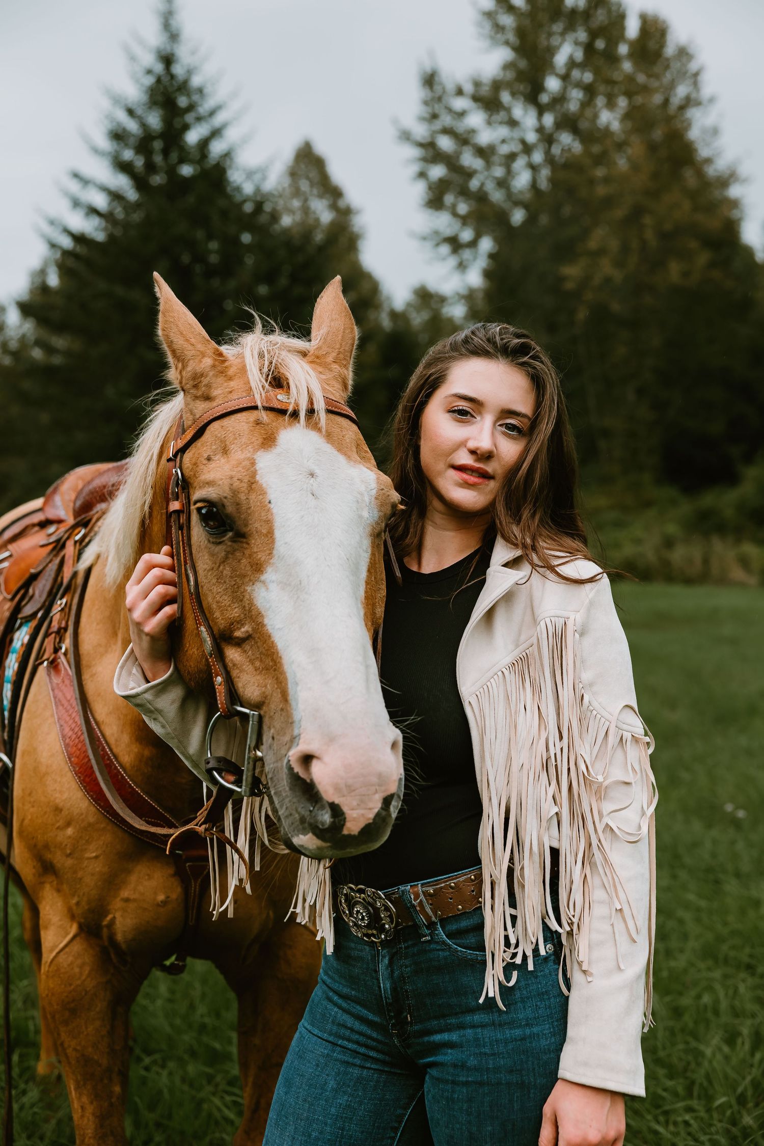 A beautiful brown and white horse with its rider wearing a fringed jacket stands in a green field on an overcast day.