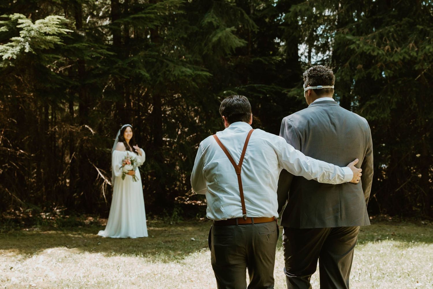 Two groomsmen watch as bride poses in forest setting for photos.
