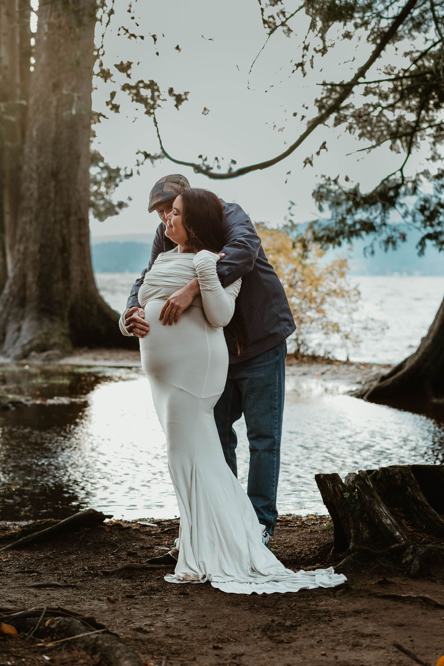 A tender embrace captured in front of tall trees with a waterfront view during a maternity session.