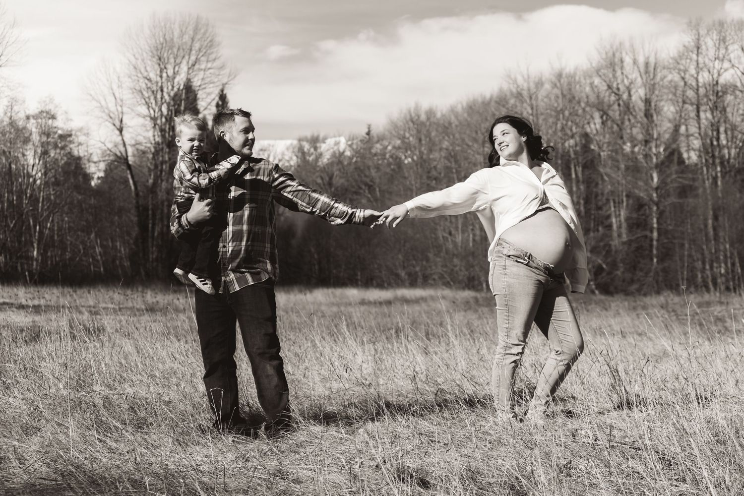 Playful couple poses in a field during maternity photo session in black and white.
