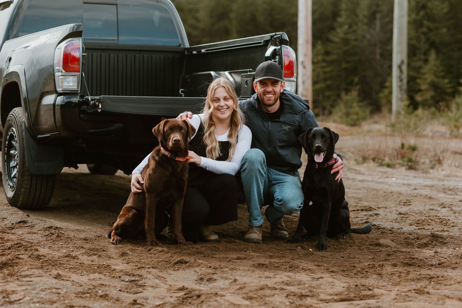 Two Labrador dogs sit next to pair of people by a pickup truck on dirt road in outdoor setting.