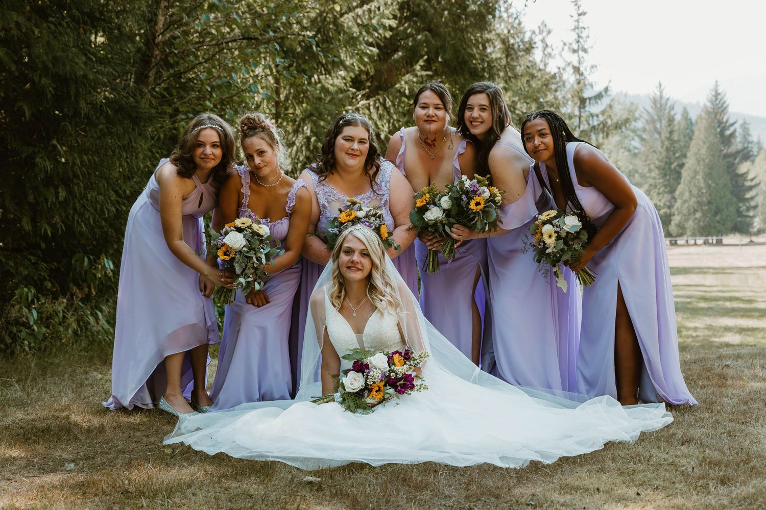 Bridesmaids in lavender dresses gather around bride for group photo outdoors.