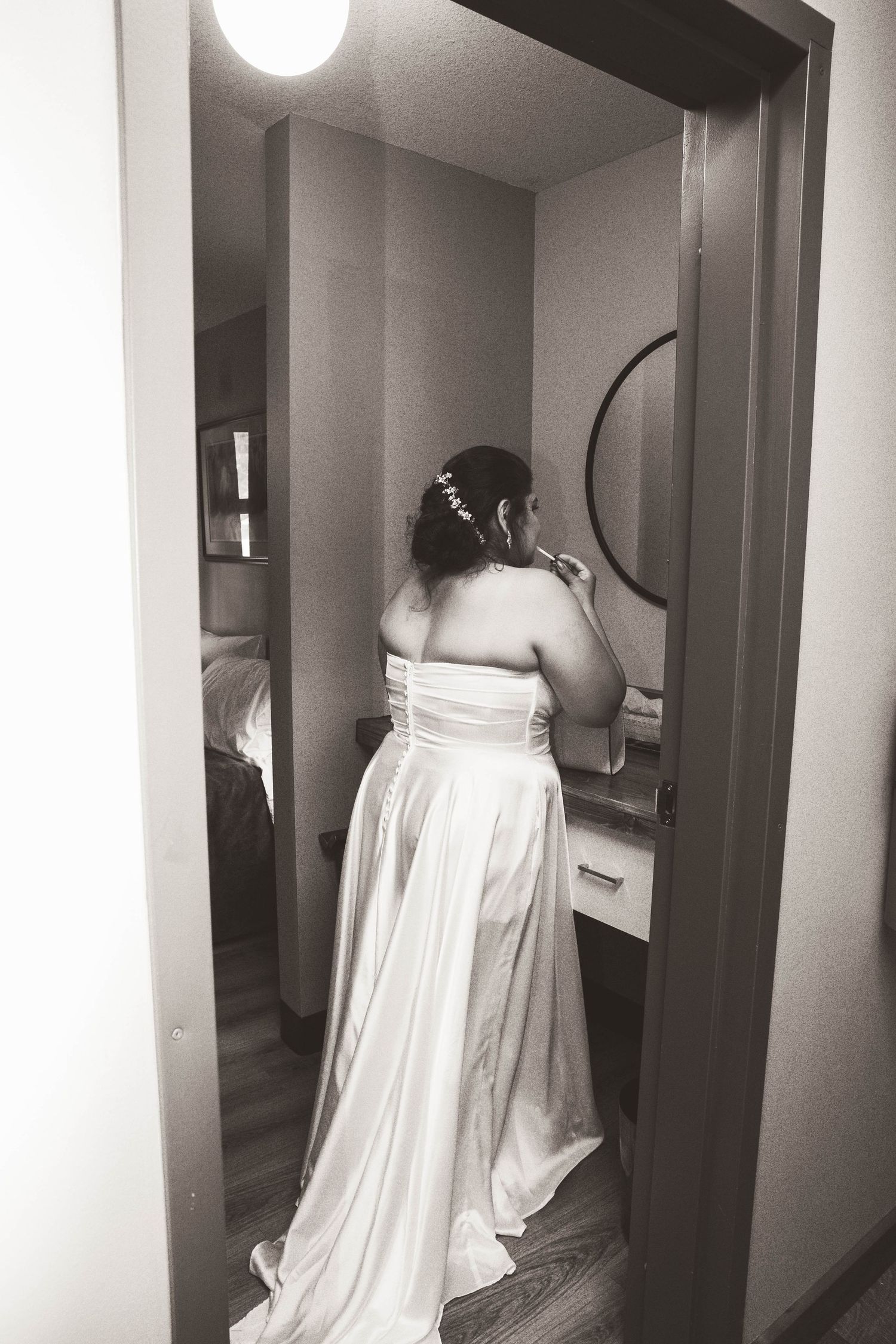 Black and white photo of a bride in her wedding dress getting ready in front of a mirror.