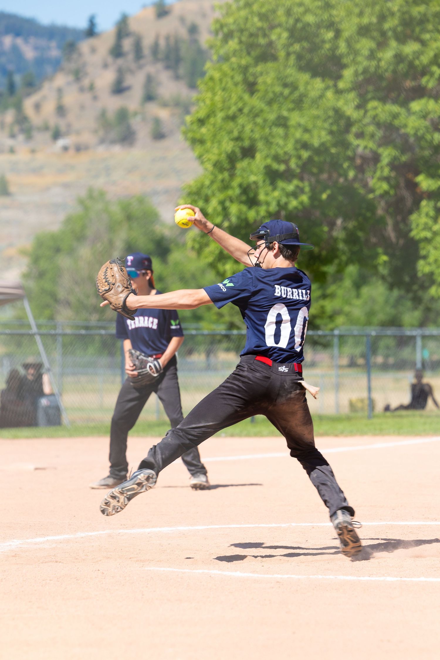A softball pitcher in mid-throw motion on a dusty diamond field with mountains in background.