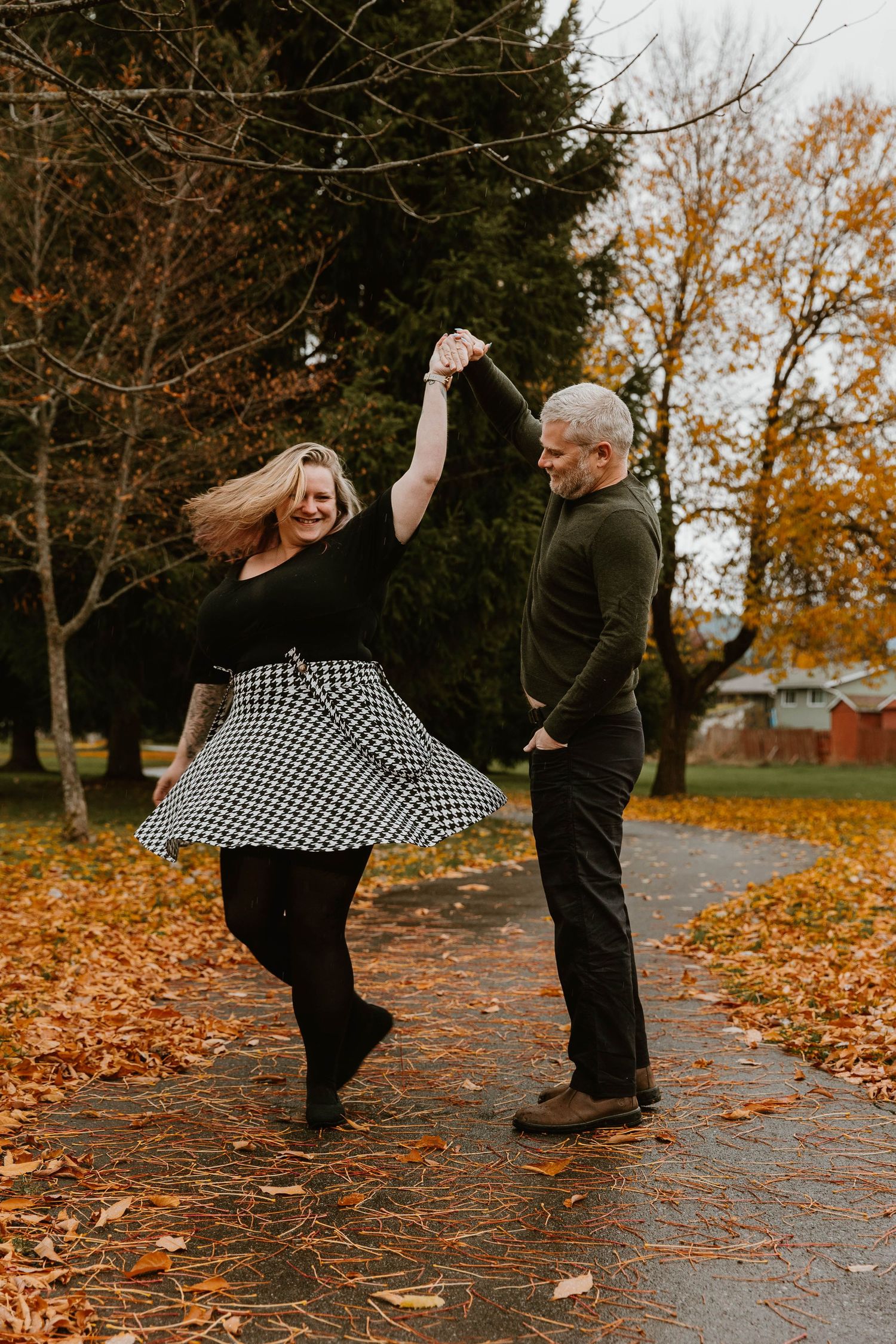 A couple dances playfully on a leaf-covered path during autumn, with the woman's checkered skirt twirling.