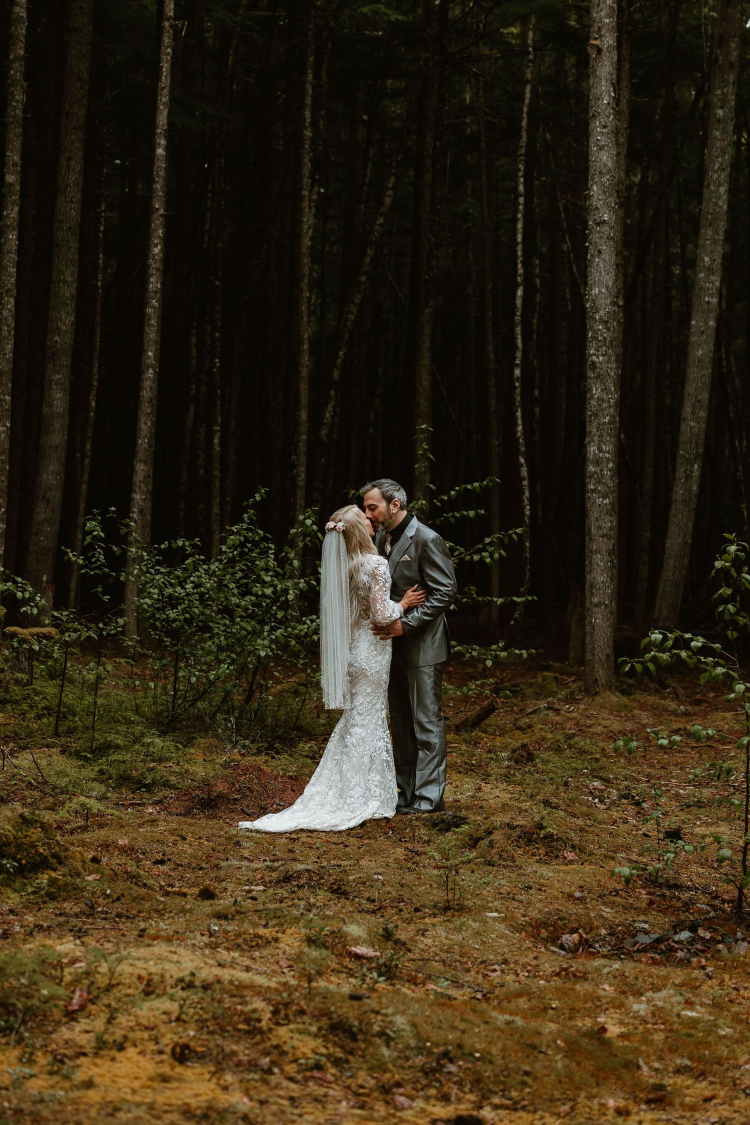 A couple shares a romantic moment in a moody forest wedding scene among tall pine trees.