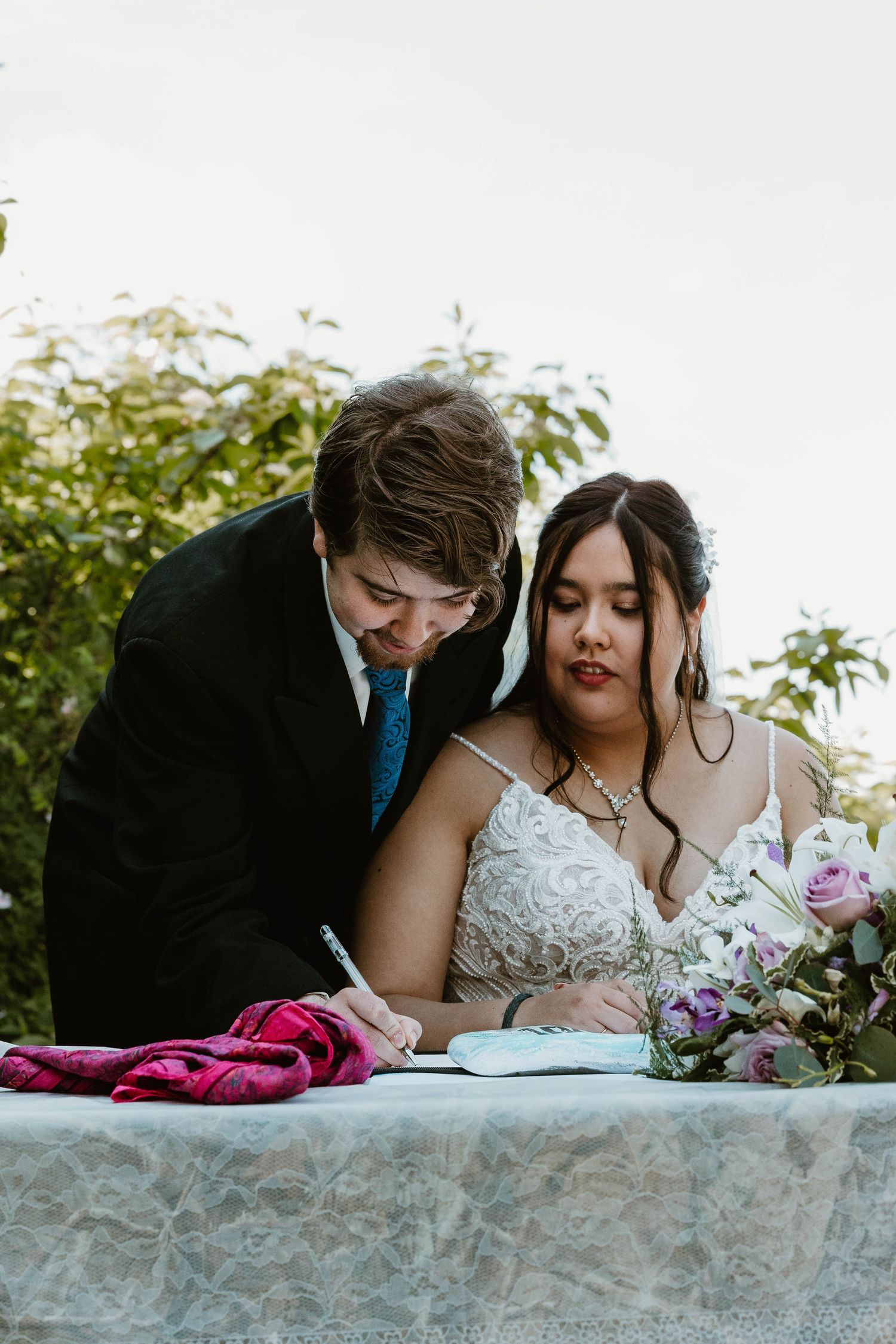 Wedding couple signing their marriage certificate at a decorated table outdoors.