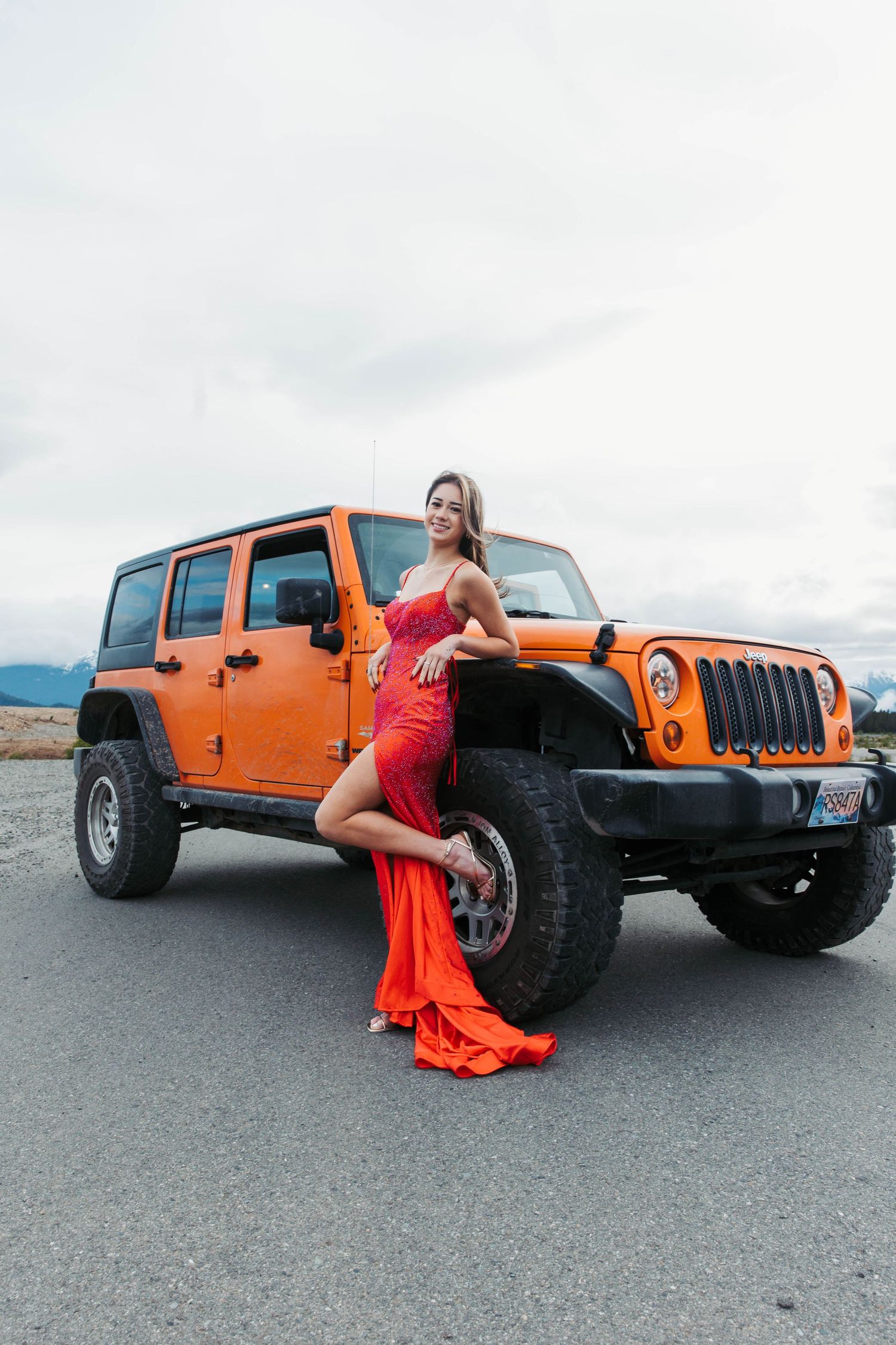 Red flowing dress contrasts with orange Jeep Wrangler against cloudy mountain backdrop.