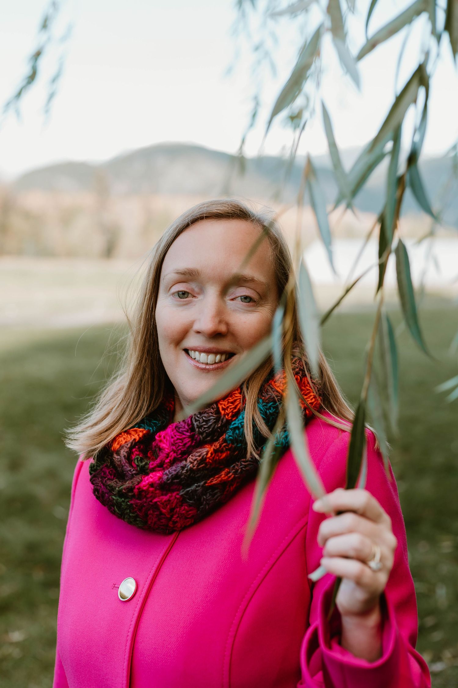 Portrait through willow branches wearing bright pink coat and patterned scarf in natural setting.