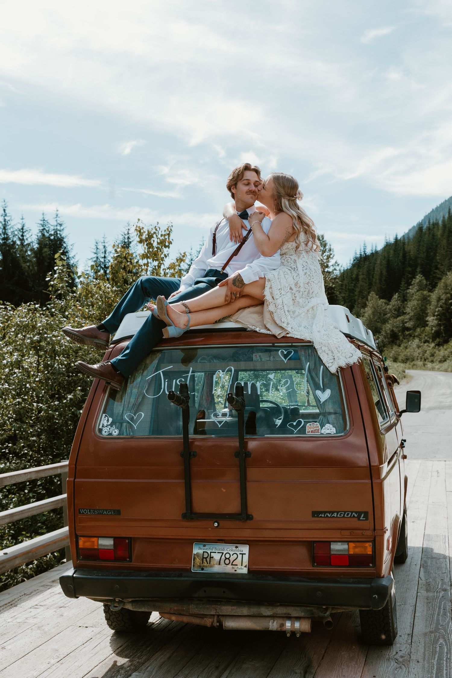 A couple sits atop an orange vintage van against a backdrop of mountain forests.