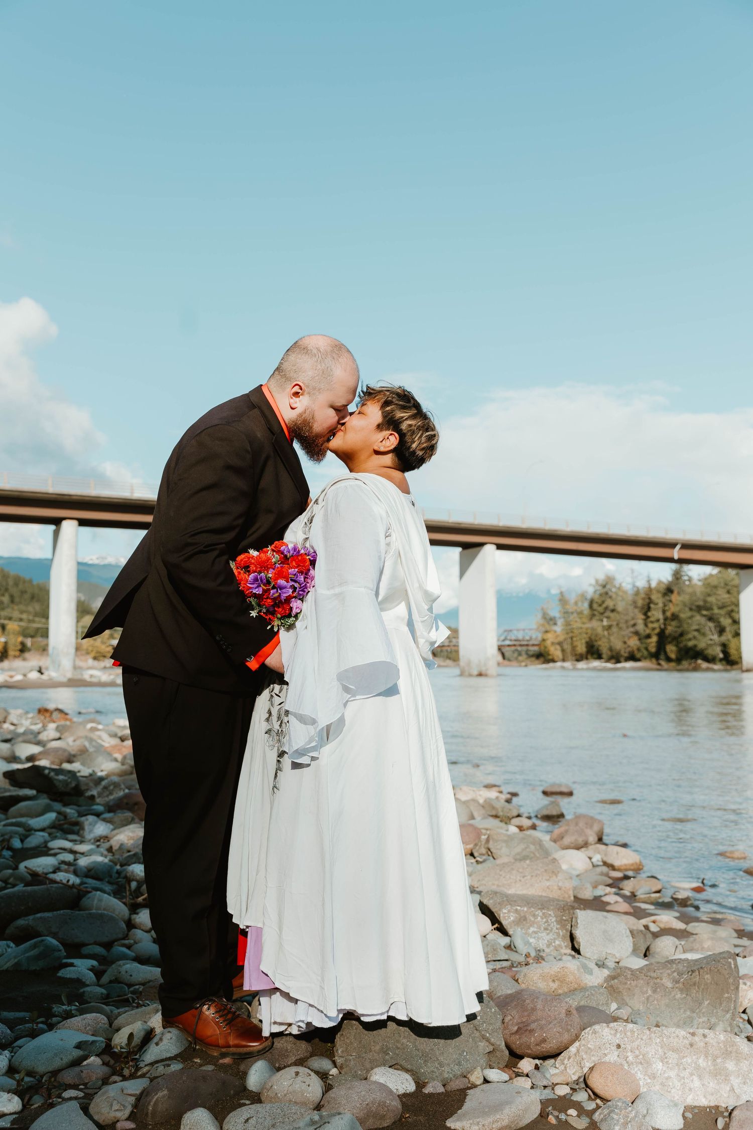 A romantic wedding couple embraces on rocky riverbank with bridge in background.