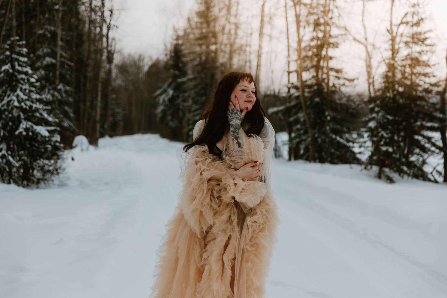 Person in flowing beige dress standing on snowy winter road surrounded by evergreen forest.