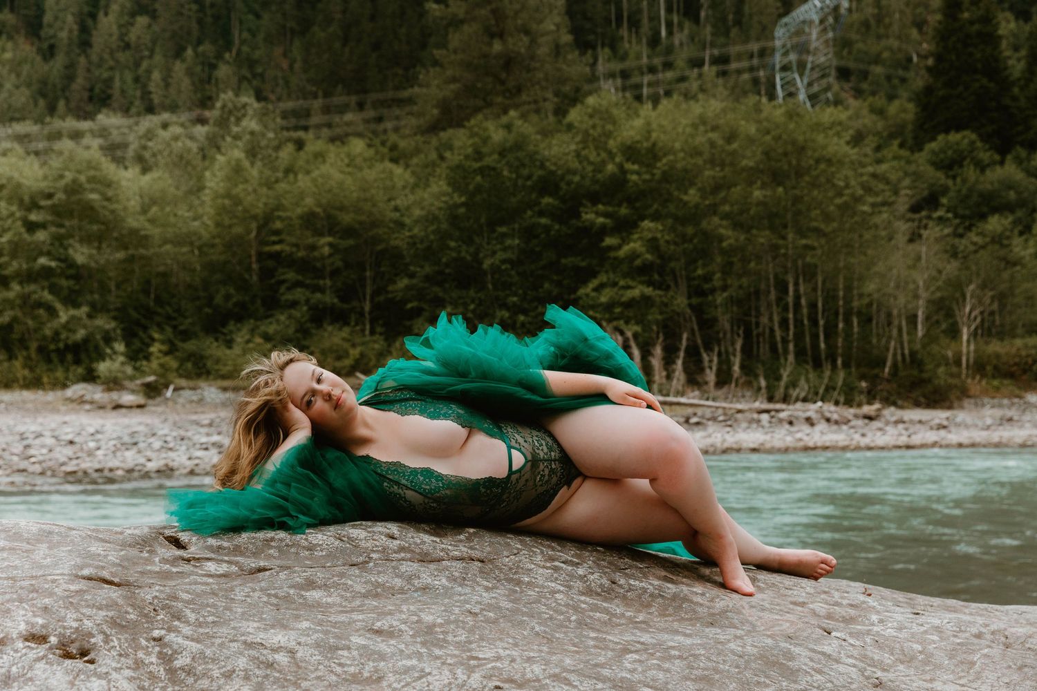 Person in emerald dress poses dramatically on rocky riverbank in forest setting.