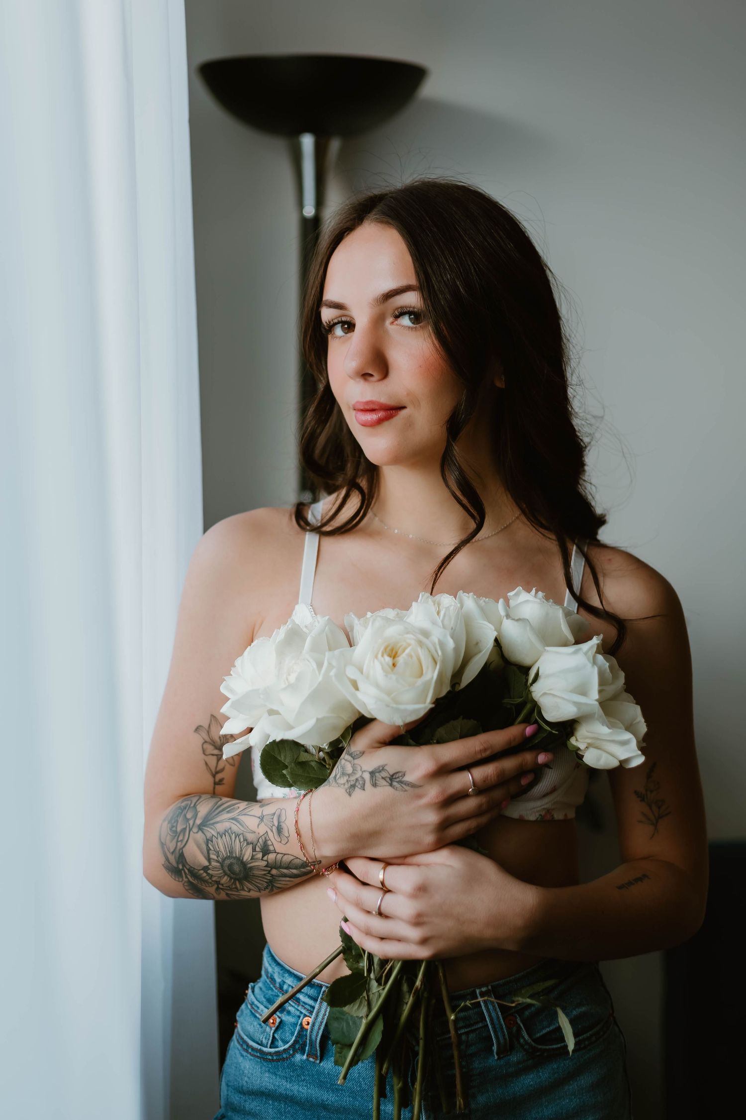 Woman holding white roses poses by window in casual denim.