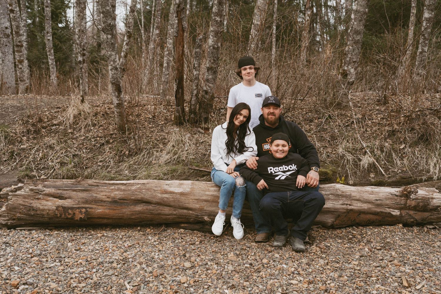 Four people sit together on a fallen log in a wooded forest area during autumn.