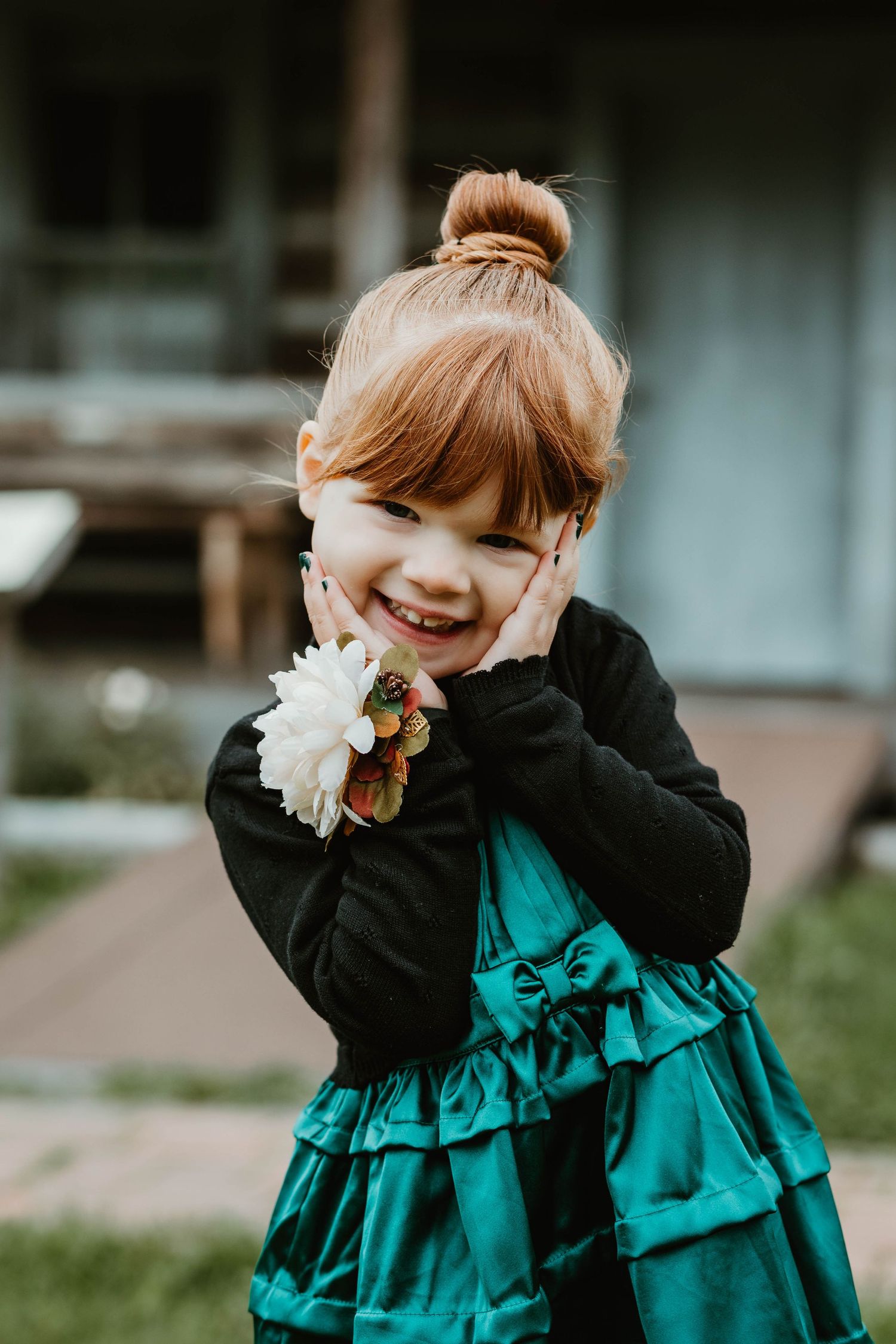 A child in a teal party dress and black cardigan giggles while holding a white flower.