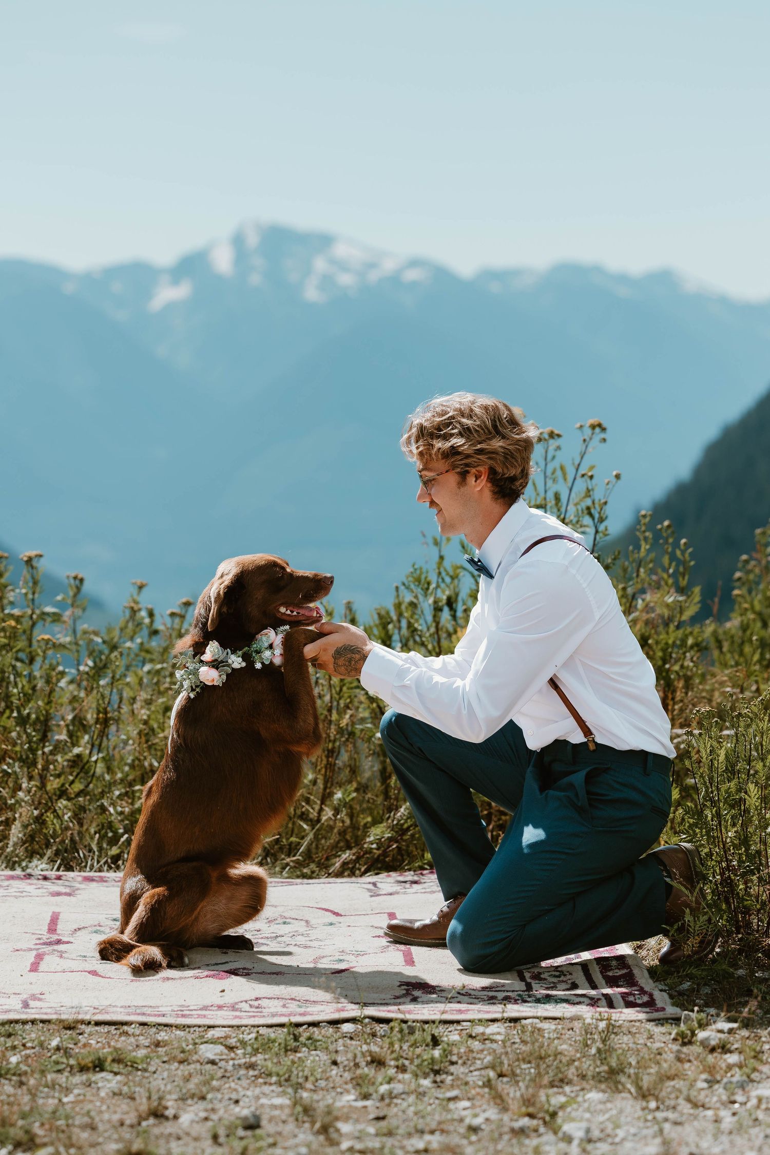 Person in formal wear kneels to interact with brown dog wearing flower crown against mountain backdrop.