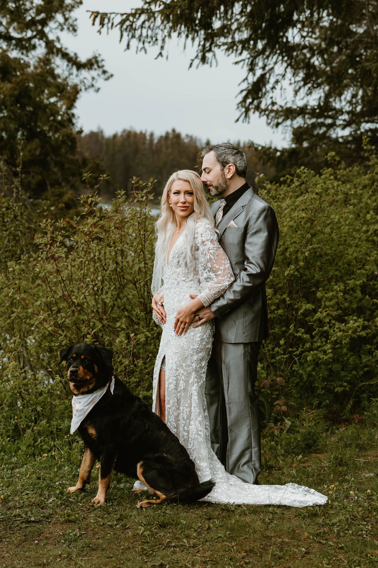 A couple in wedding attire enjoying an intimate outdoor moment in a natural setting with their dog nearby.