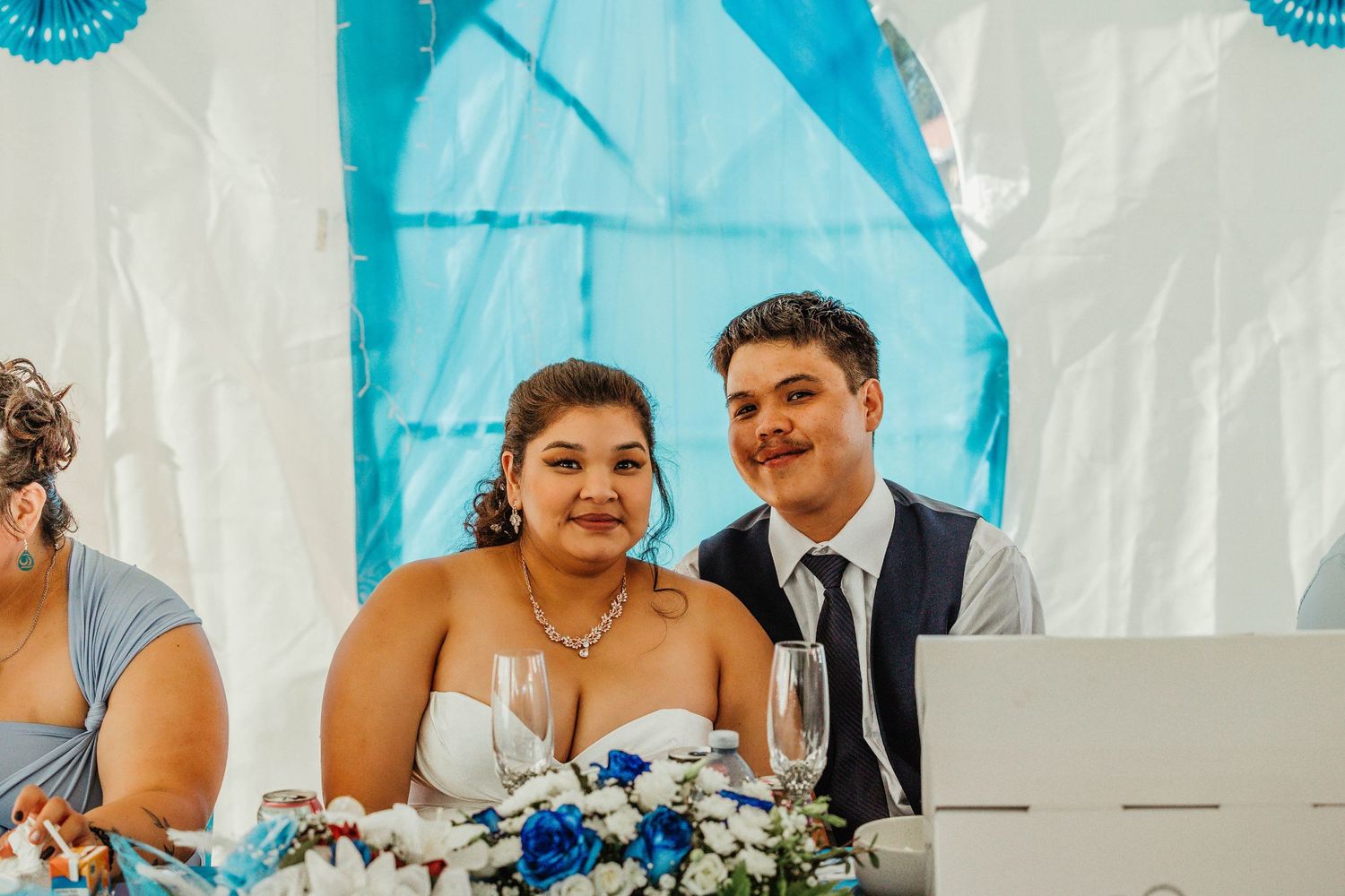 Newlyweds sitting at reception table decorated with blue accents and white flowers.