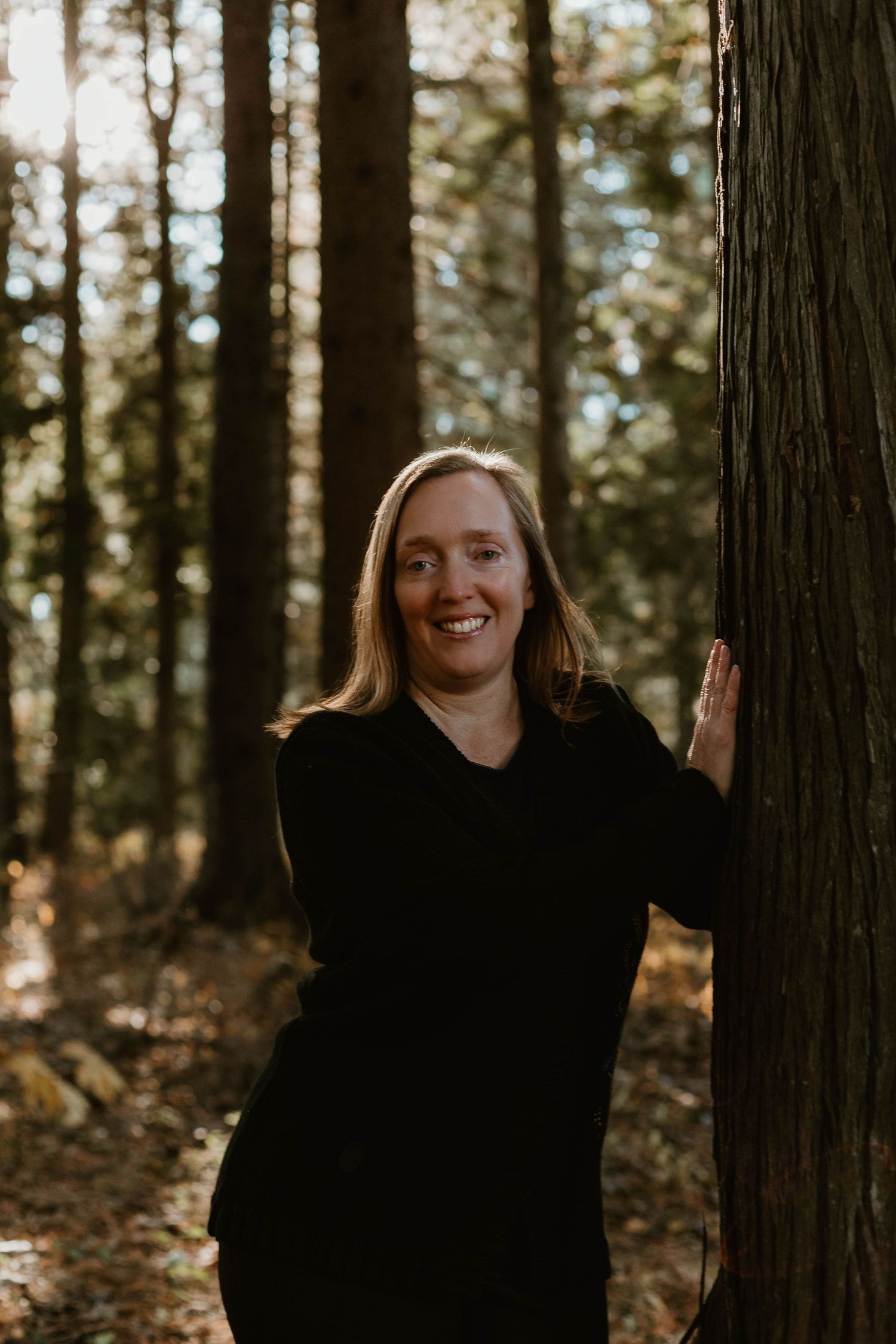 Candid forest portrait in dark clothing leaning against tall evergreen tree.