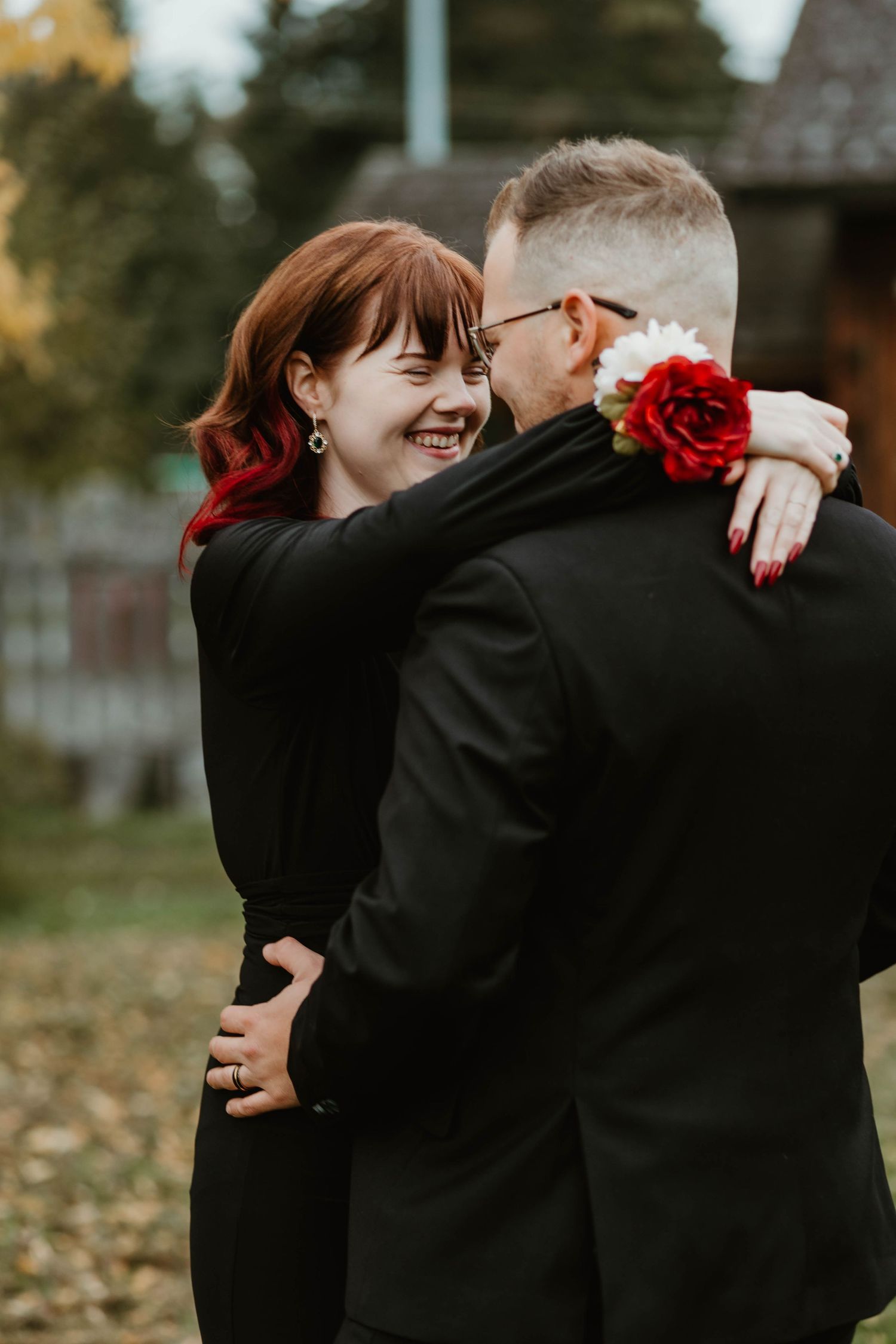 A couple embraces in formal black attire while sharing a romantic moment outdoors.