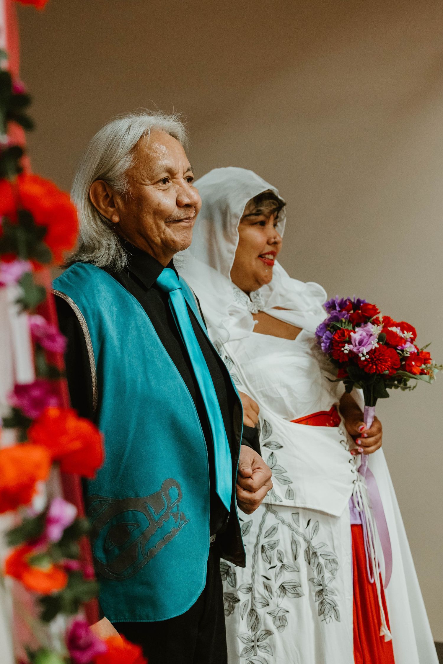 Bride in white dress holds colorful bouquet while standing with guest in turquoise jacket.