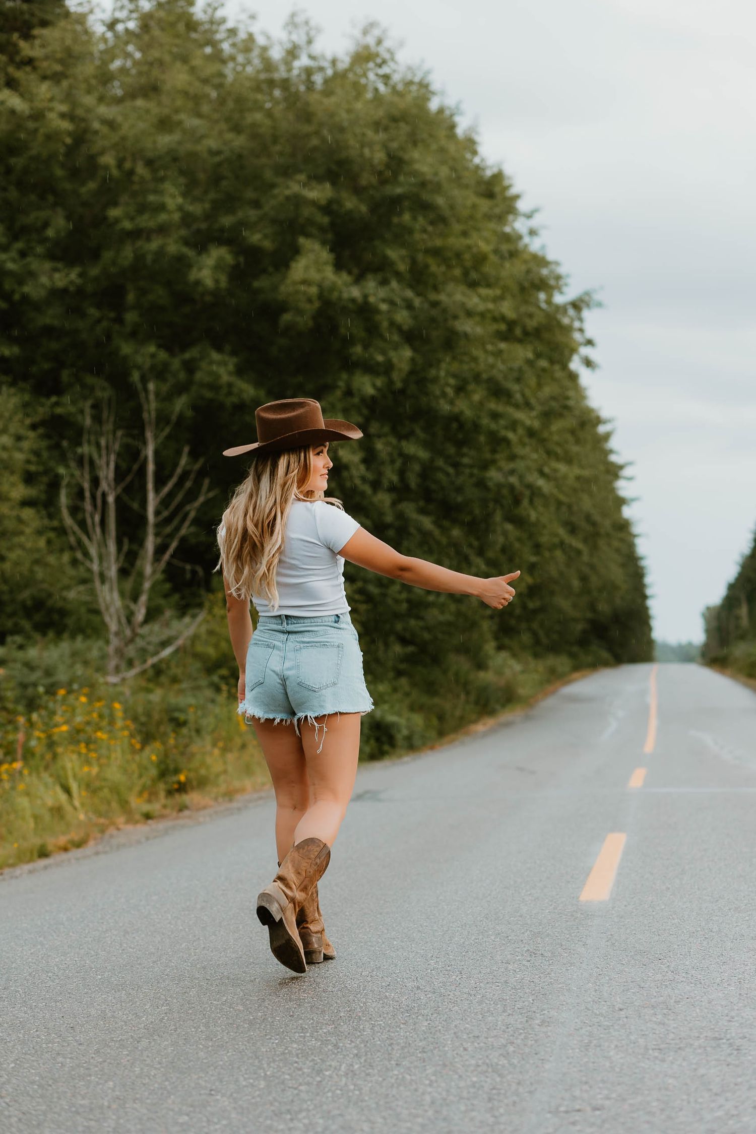 Girl in denim shorts and cowboy hat walking down empty rural road.