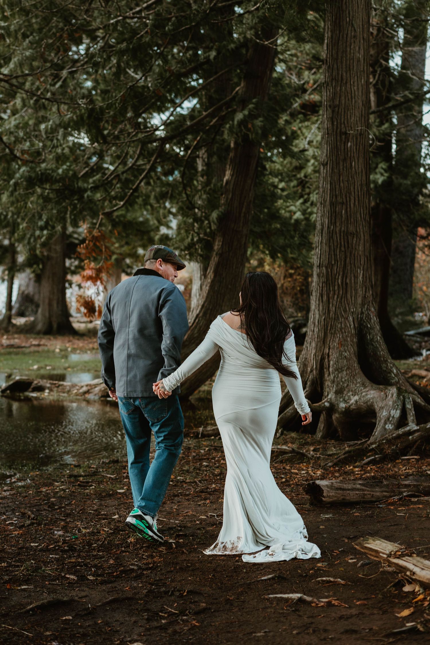 A couple walks hand in hand through a wooded area wearing formal and casual attire.