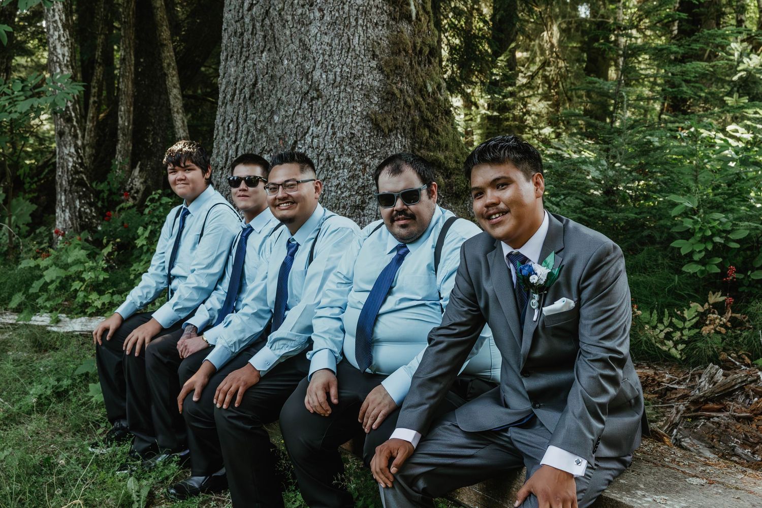 Groomsmen in light blue shirts and grey suits seated on a log in a forest setting.