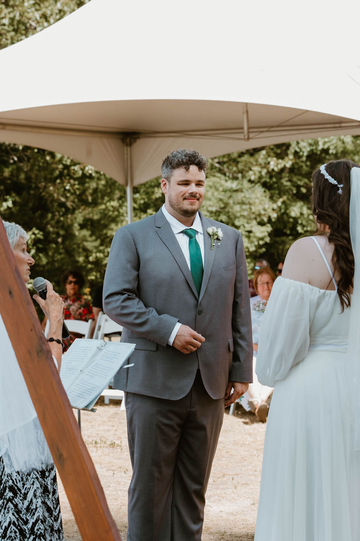 An outdoor wedding ceremony features a couple exchanging vows under a white canopy with wooden decor.