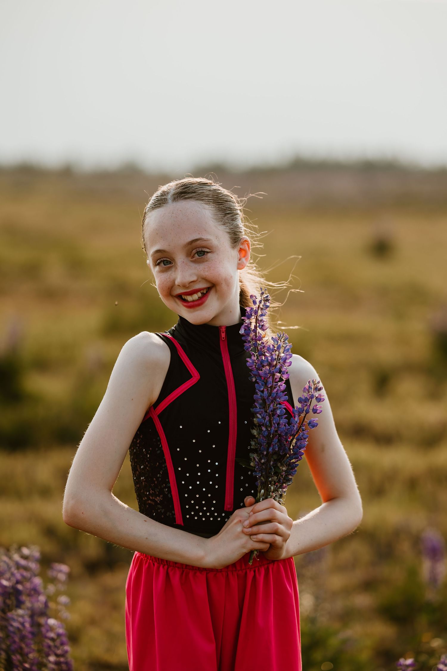 A red-dressed figure dances joyfully in a natural field at golden hour, holding purple wildflowers.