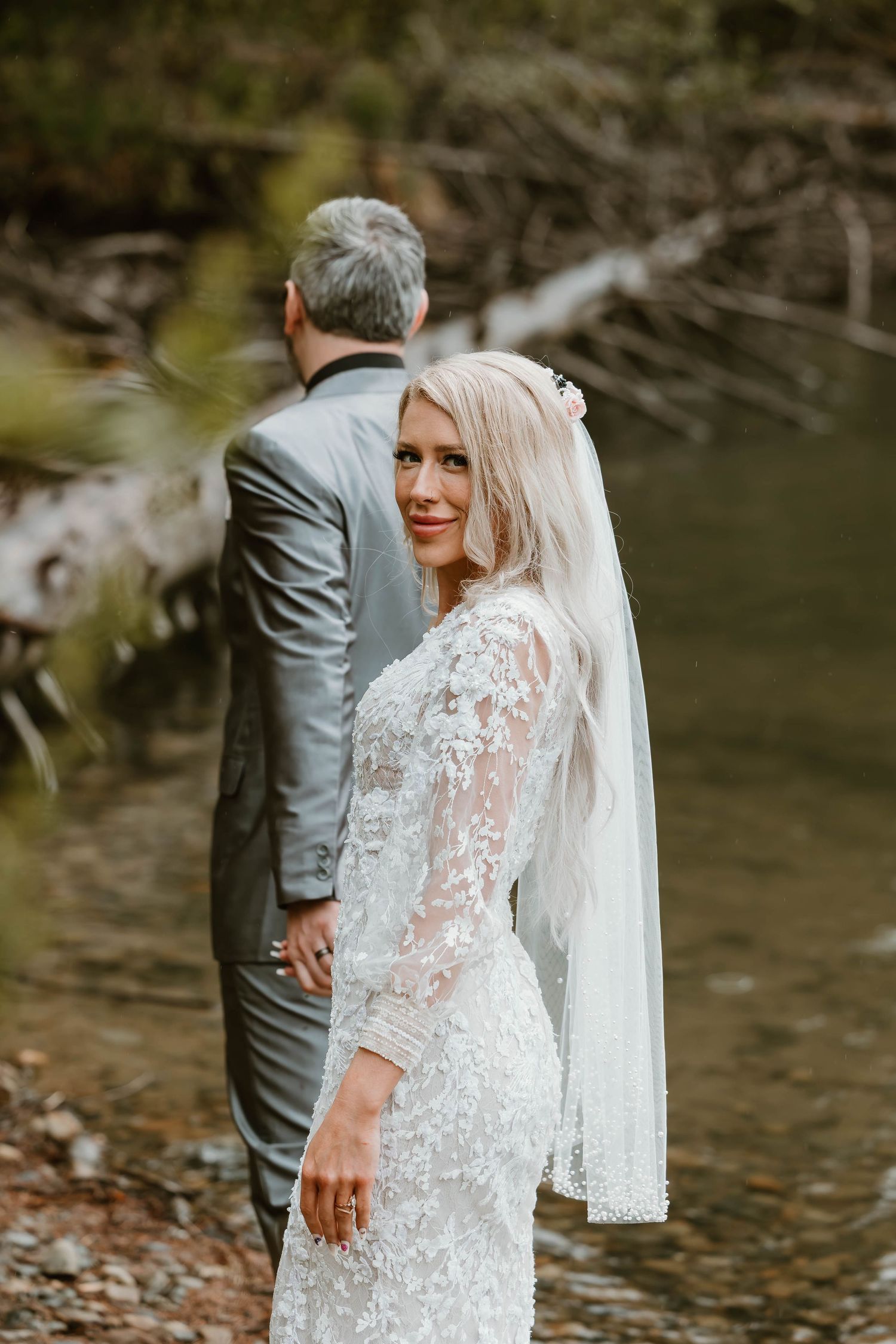 A couple shares a romantic first look moment by a creek, with the bride wearing an elegant lace long-sleeved wedding gown.