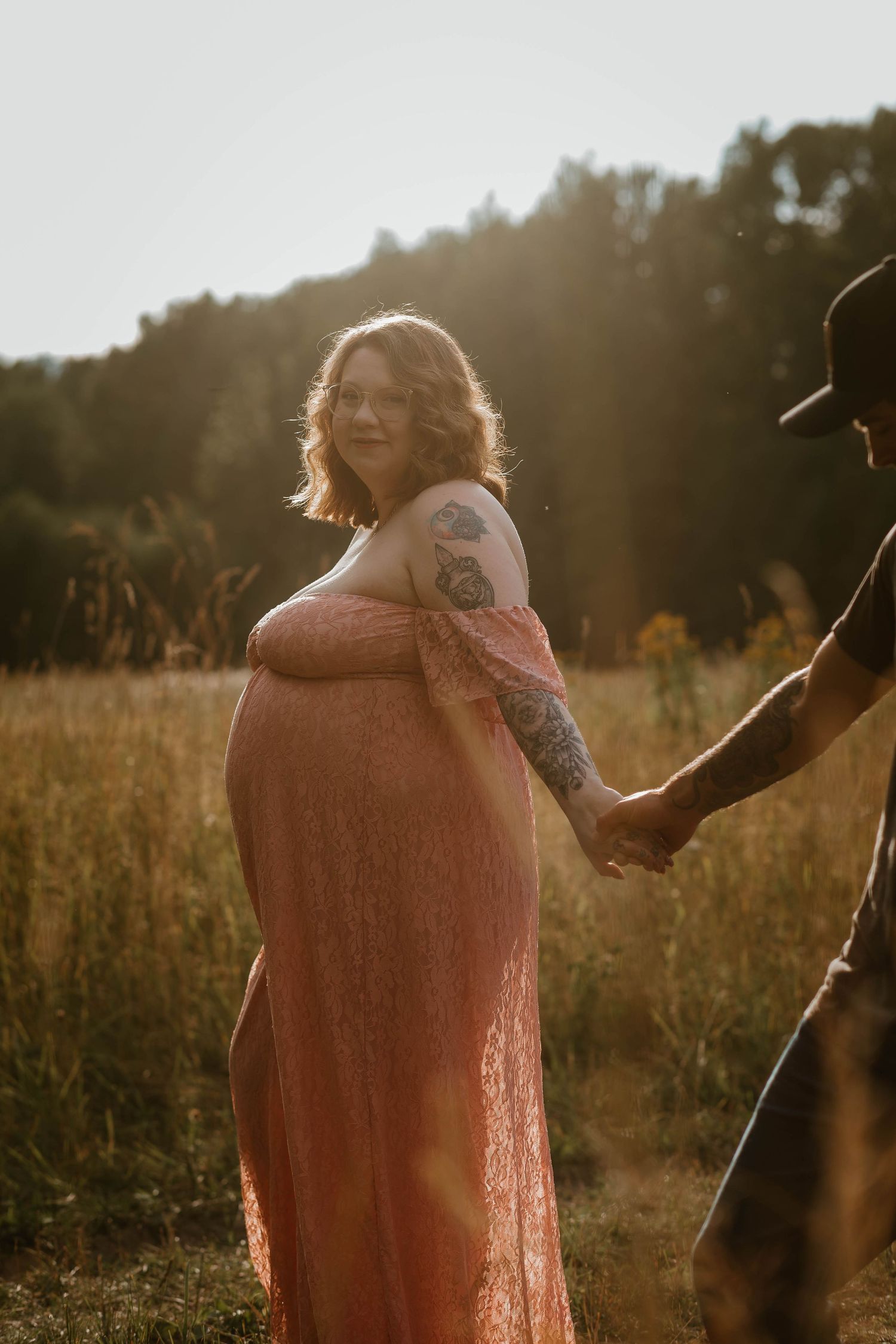 A pregnant woman in a pink dress stands in a golden sunlit field.