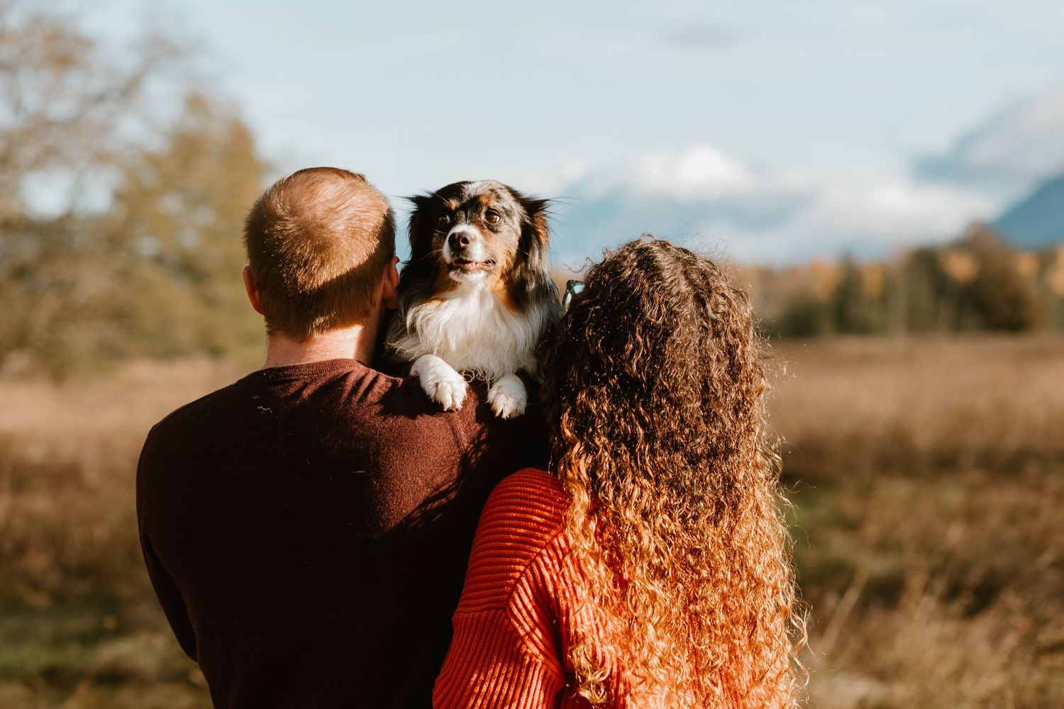 A couple holds their Australian Shepherd dog while enjoying an autumn day in nature with mountains in background.