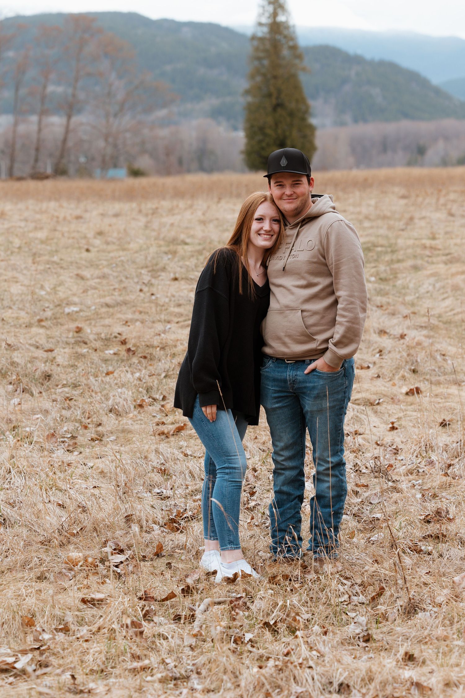 Couple poses together in autumn field with mountains and evergreen trees in background.